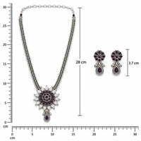 A traditional Indian jewelry set featuring a silver-toned necklace with a floral pendant, matching drop earrings, and a beaded chain, decorated with dark purple stones and clear crystals, displayed on a light pink background.